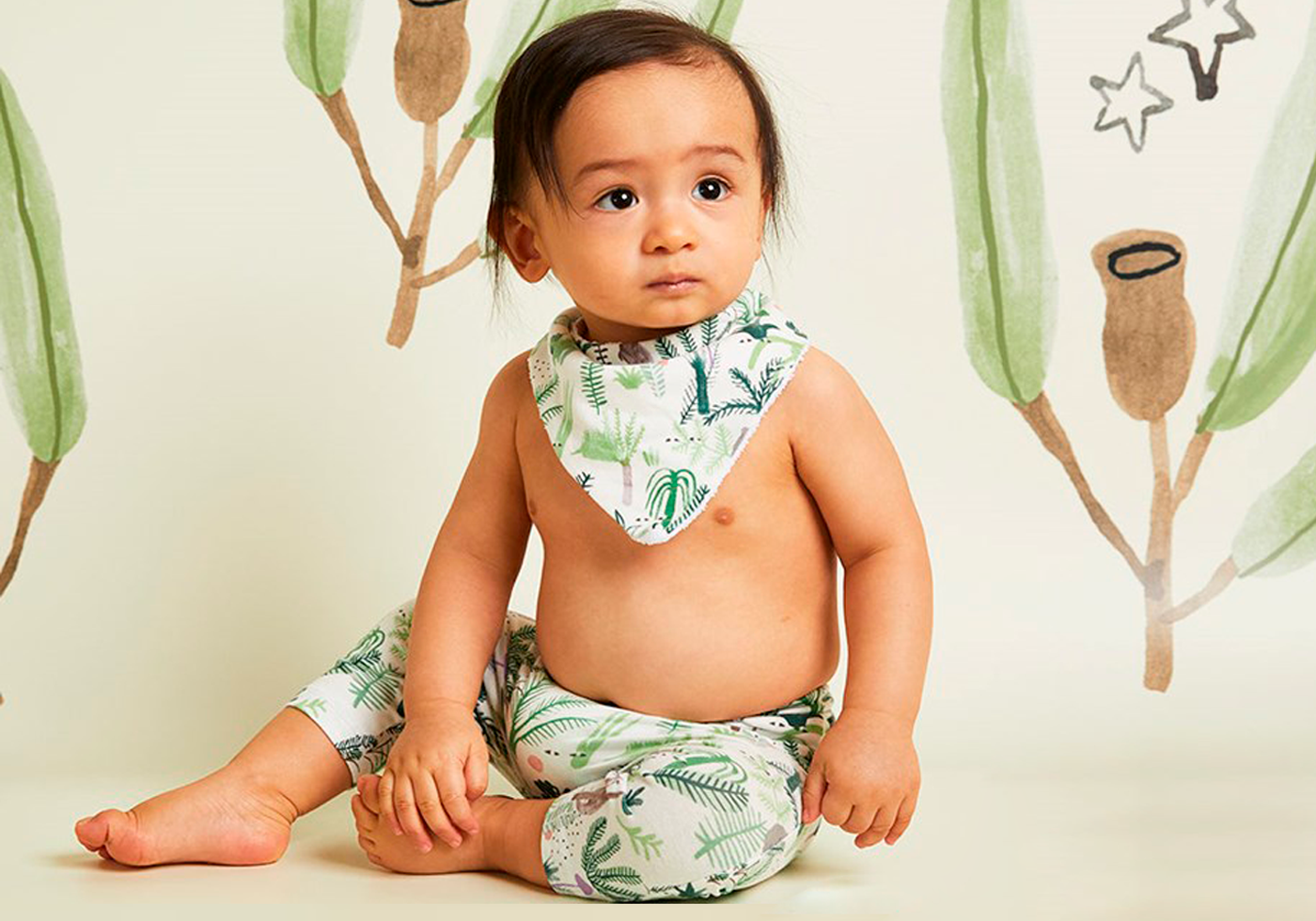 Baby wearing a green leaf-patterned outfit sitting on a beige surface with decorative elements in the background.