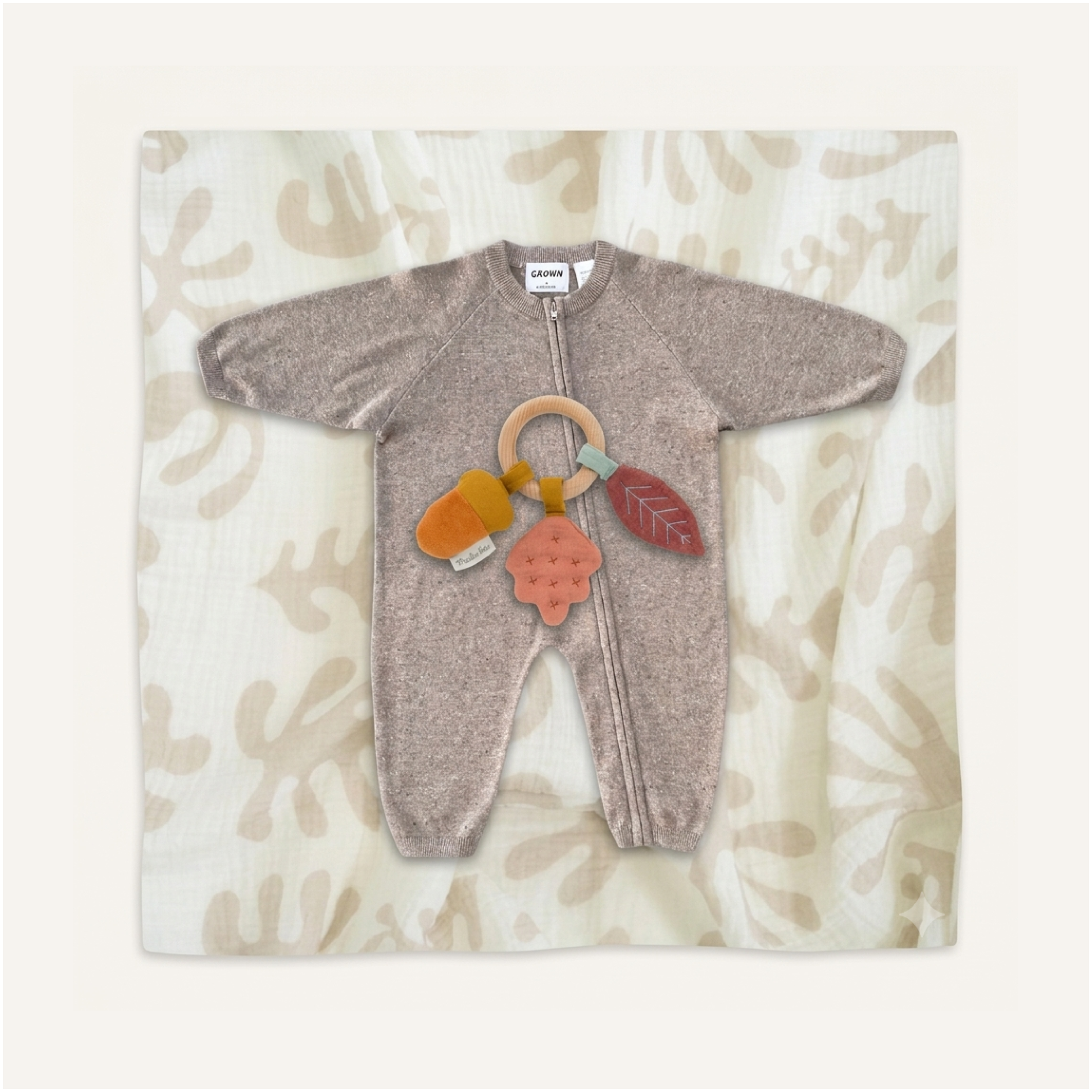 Golden Beginnings Baby Set by Clay and Hutch. Baby clothing set by Grown, Golden Child and Moulin Roty. A baby sleep sack in a heathered taupe-brown color, laid flat on a cream-colored muslin blanket with a beige leaf pattern. The sleep sack is styled with a wooden baby teething ring and three fabric teething toys in orange, pink, and red.